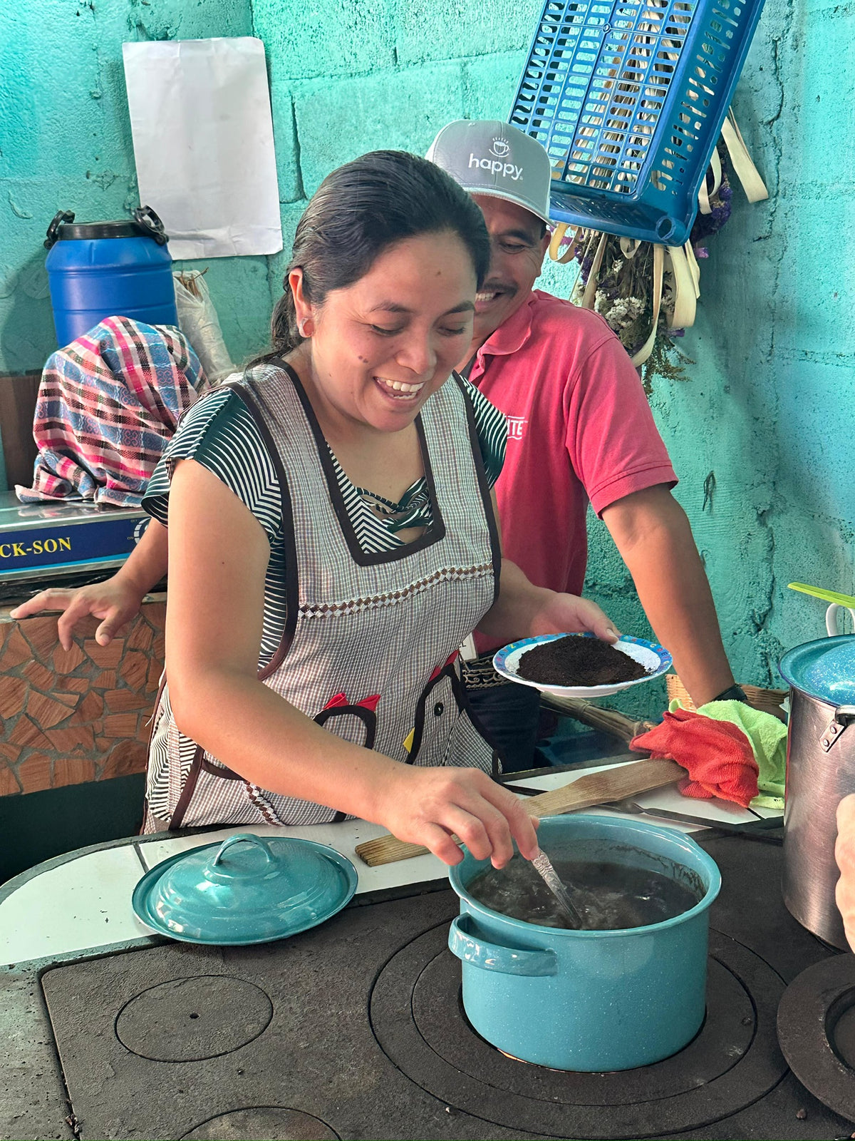 A woman in a kitchen wearing a black and white striped apron over a patterned top, stirring a pot on a rustic stove while holding a plate. A man in a pink shirt stands behind her.