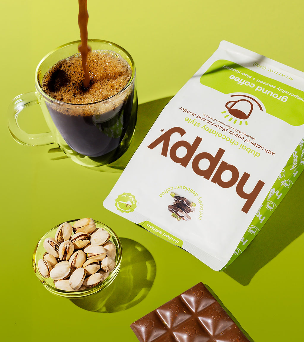 Glass Mug of coffee, bowl of Pistachios, Dubai Chocolate Bar and package of Happy Dubai Chocolatey Style Ground Coffee on a green background