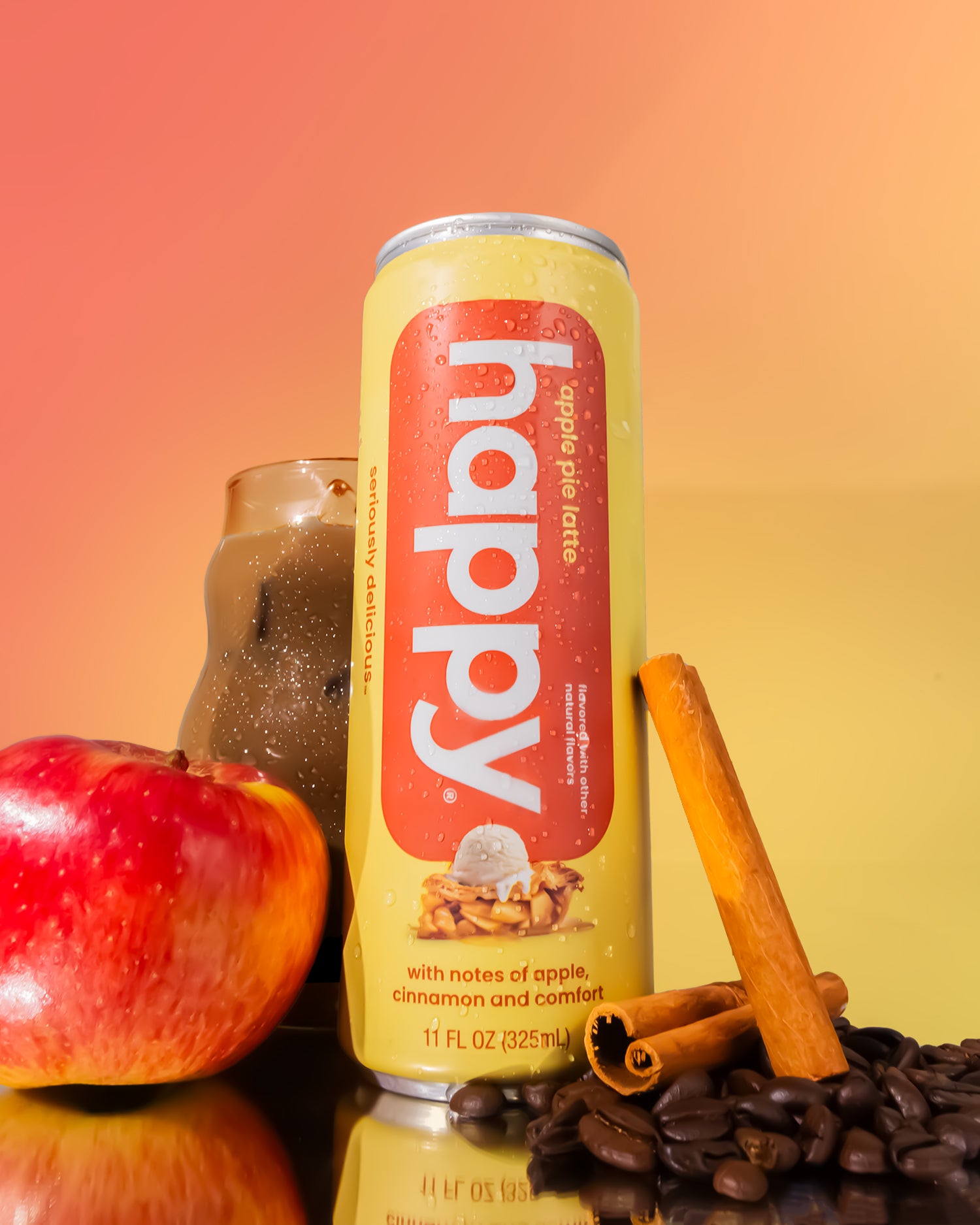Can of 'happy' apple pie latte with an apple, cinnamon stick, and coffee beans on a gradient background