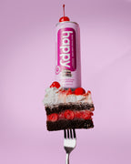 Pink can of Happy Black Forest Cake Latte sitting on top of piece of chocolate cake with whipped cream resting on a fork.  
