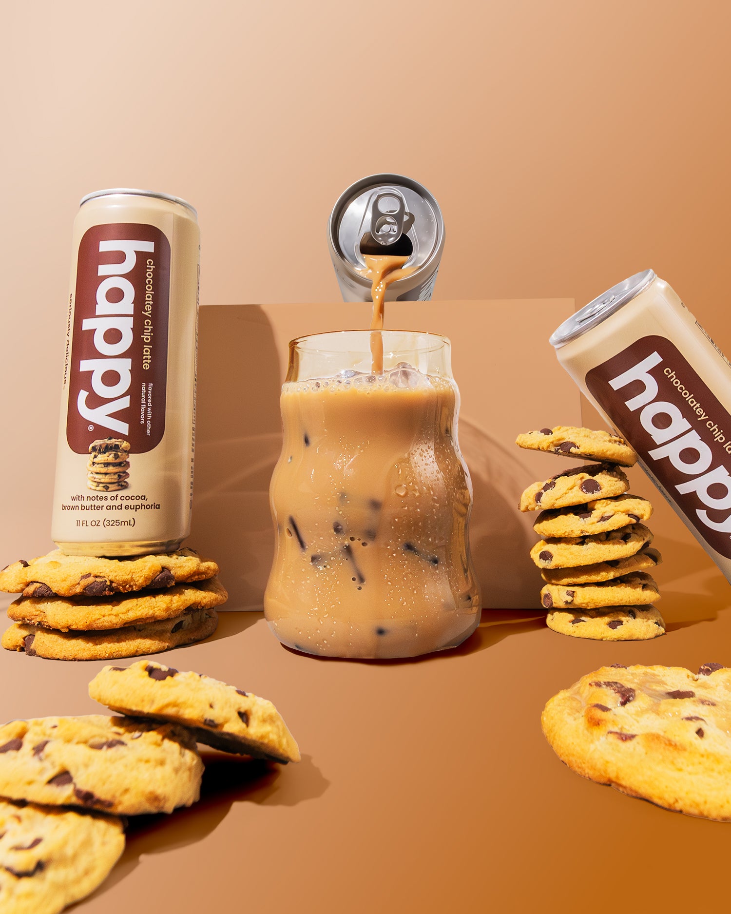 Cans of 'happy' chocolatey chip latte with a glass of iced coffee and cookies on a brown background