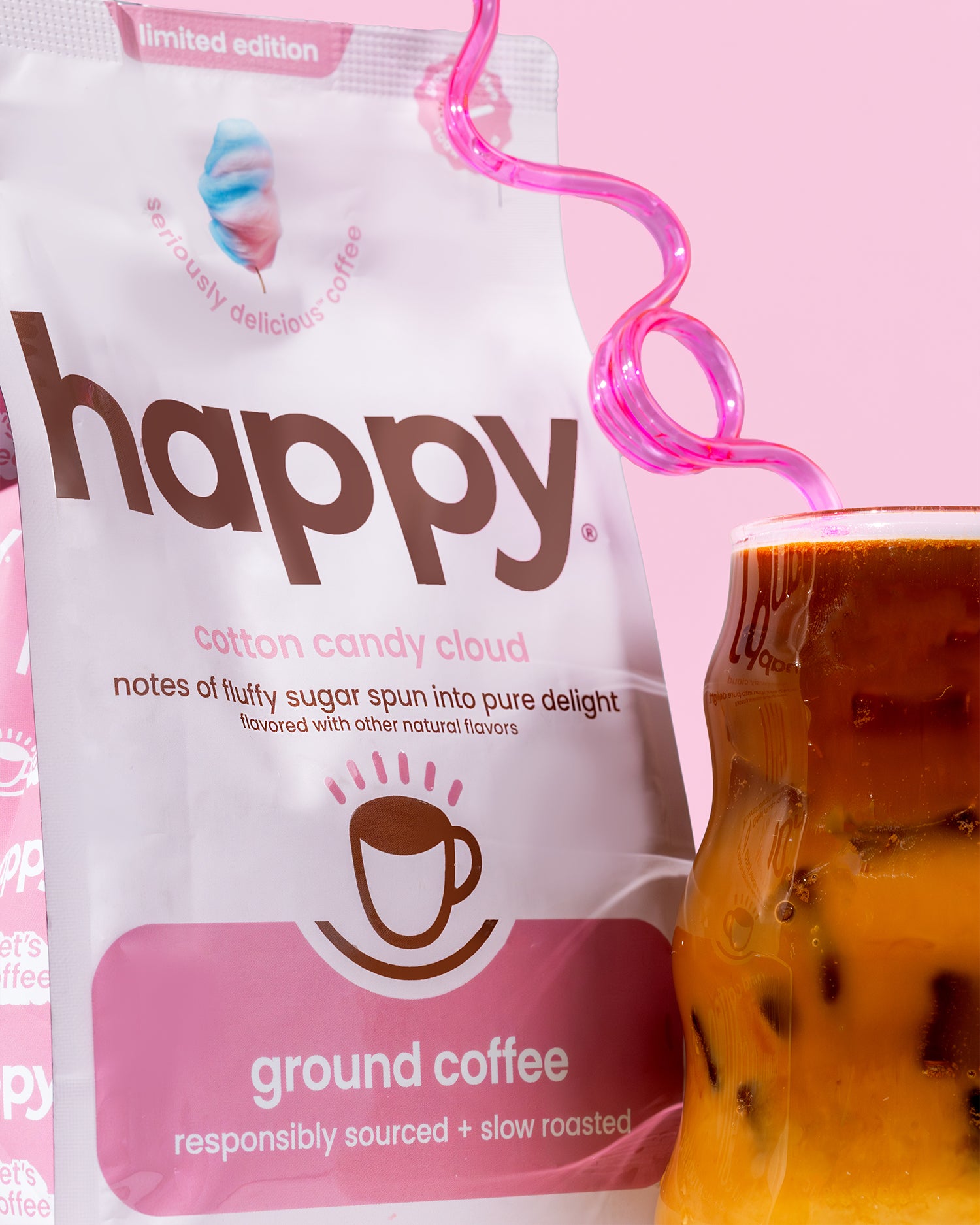 Happy cotton candy cloud ground coffee package with a pink and white design next to a cup of coffee with a pink straw.
