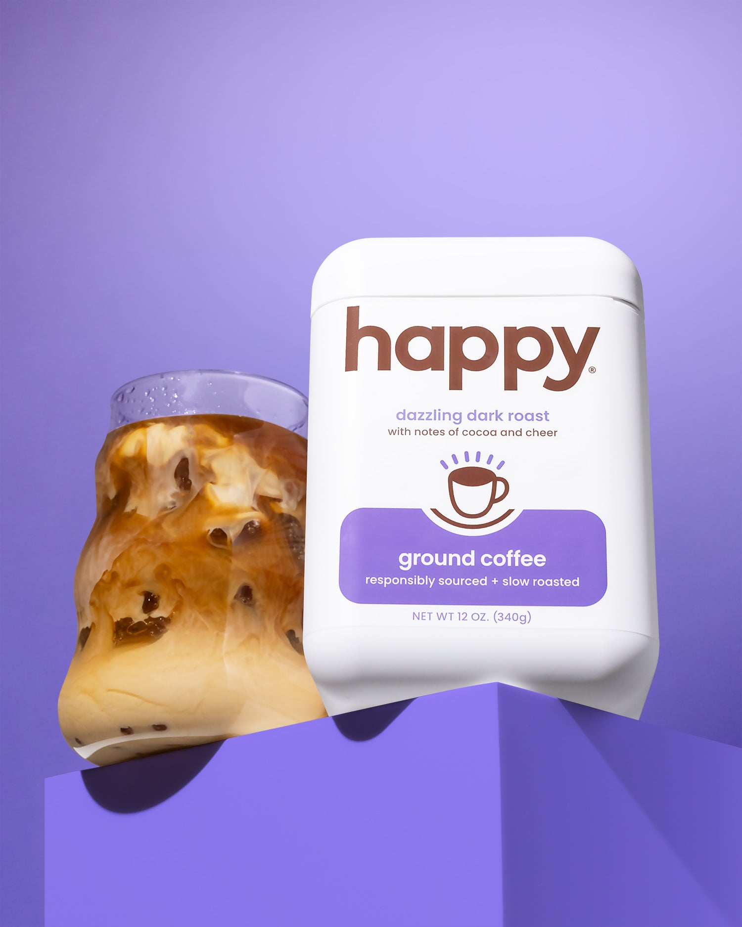 White container of 'happy' brand ground coffee next to a glass of iced coffee on a purple background