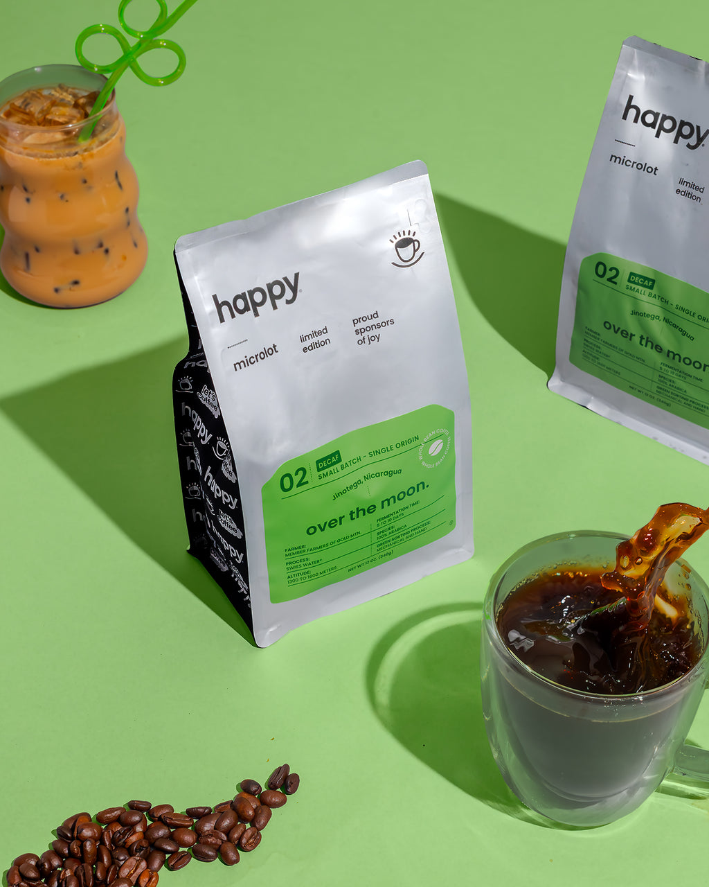 Over the Moon Decaf Coffee bag from happy on a green background with coffee beans, a glass of coffee, and a latte with a cute green straw.