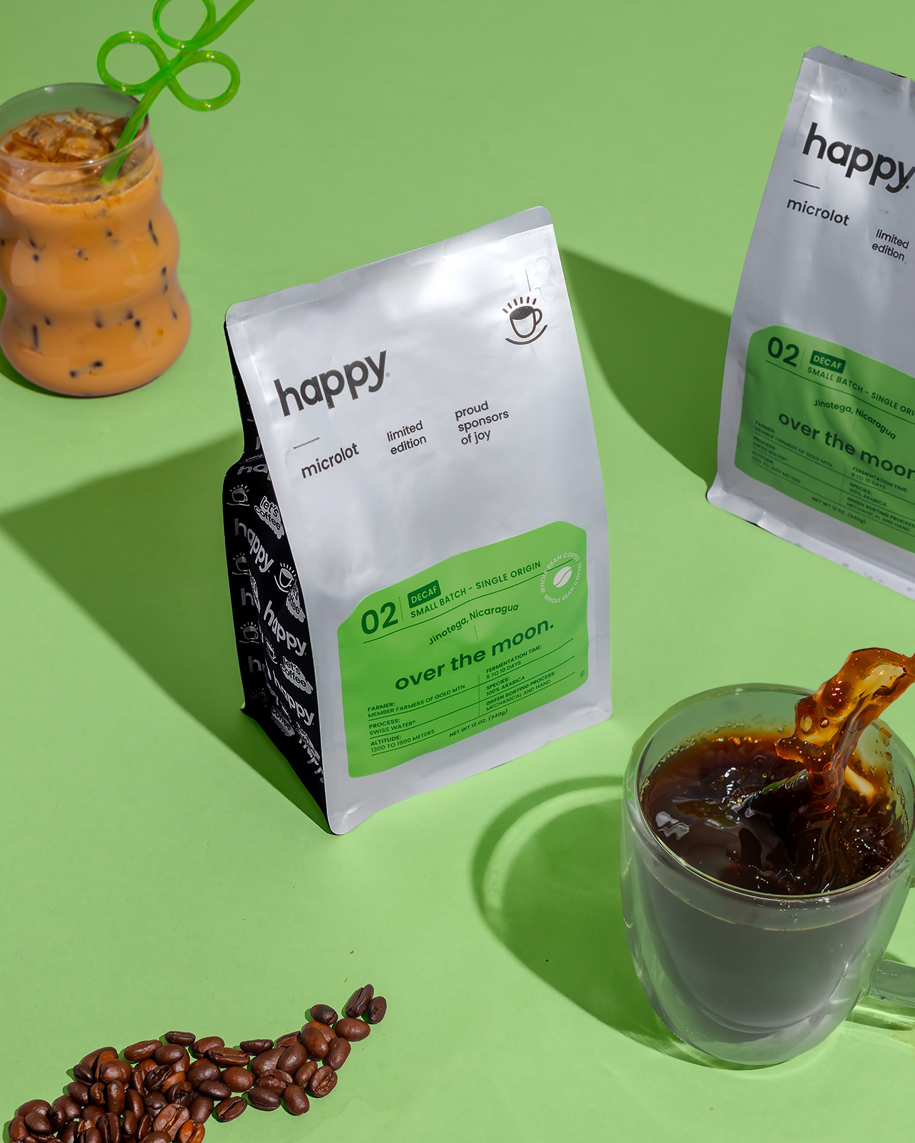 Over the Moon Decaf Coffee bag from happy on a green background with coffee beans, a glass of coffee, and a latte with a cute green straw.