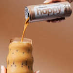 Person pouring a mocha latte from happy coffee into a glass against a warm brown background.