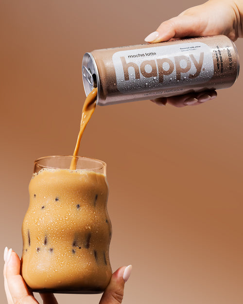 Person pouring a mocha latte from happy coffee into a glass against a warm brown background.