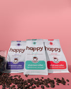 Three 'happy' brand whole bean coffee packages with scattered coffee beans on a pink background