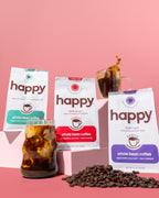 Three packages of 'happy' whole bean coffee with iced coffee and coffee beans on a pink background