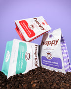 Three 'happy' brand coffee packages on a bed of coffee beans with a purple background