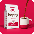 A bag of Happy Daily Magic ground coffee stands next to a white ceramic mug with a red base, with coffee being poured into the mug, set against a vibrant pink background.