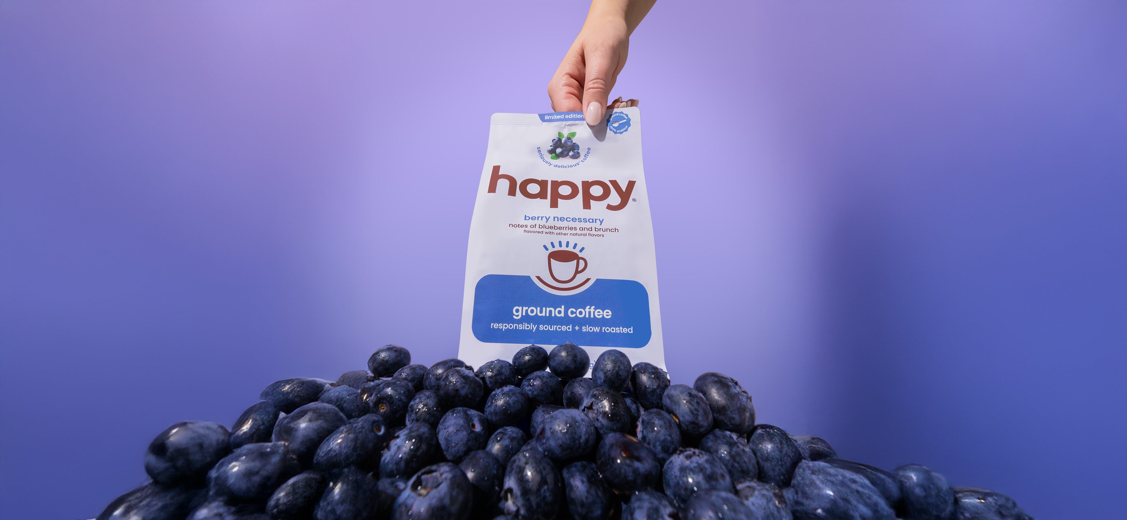 Hand holding a 'happy' coffee package over a pile of blueberries against a purple background
