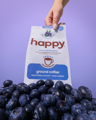 Hand holding a 'happy' brand ground coffee package with blueberries on a purple background