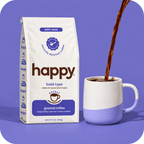 A bag of Happy Bold Type dark roast ground coffee stands next to a white and lavender ceramic mug with coffee being poured into it, set against a solid purple background.