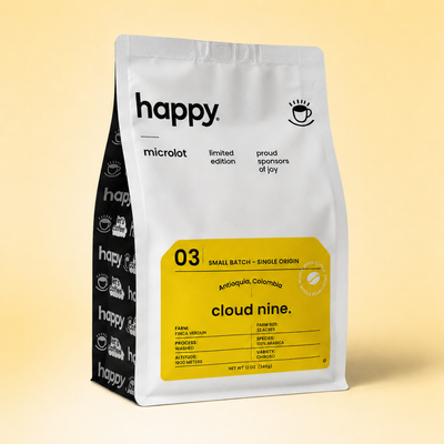 A stand-up coffee bag from Happy Microlot, featuring a silver front with a large yellow label and a black side panel with repeated coffee cup logos, photographed on a warm beige gradient background.